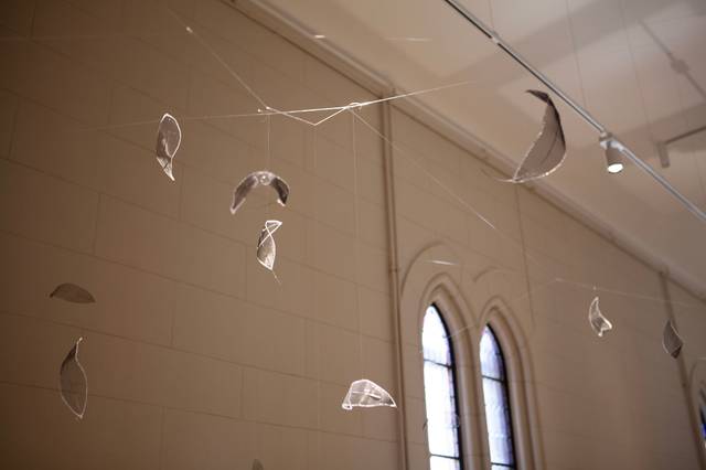 La imagen muestra una instalación artística con hojas de vidrio suspendidas en un espacio interior.