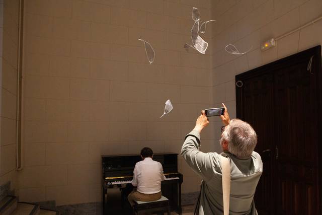 Una persona está tocando el piano mientras otra lo observa y toma una foto de objetos que flotan en el aire.