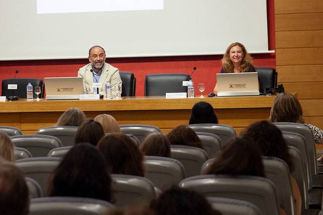 La imagen muestra a dos presentadores en un evento, sentados frente a un público en un salón de conferencias.