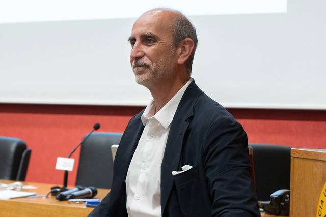 Un hombre con una chaqueta oscura y camisa blanca se encuentra sentado al frente de una sala de conferencias.
