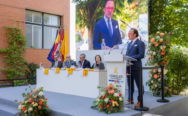 La imagen muestra un acto académico en el que un orador se dirige a un público sentado frente a él, con banderas y un gran cartel en el fondo.