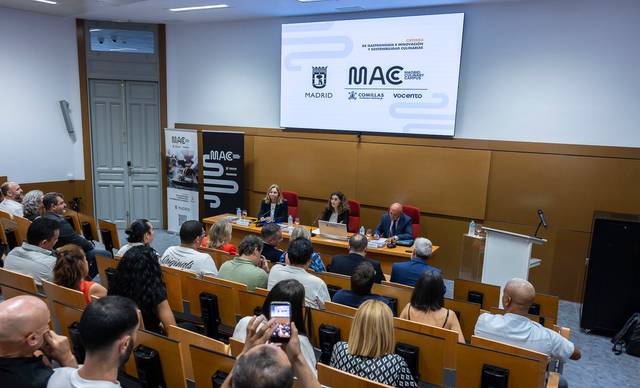 Un evento en un auditorio con varias personas sentadas y ponentes en una mesa frente a una pantalla.
