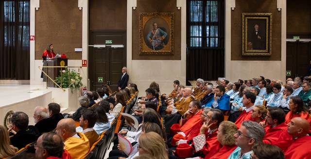 Una ceremonia académica con un orador en el podio y un público atento en un salón decorado.