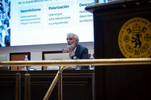 Un hombre con barba y cabello canoso se encuentra sentado frente a una presentación en una conferencia.