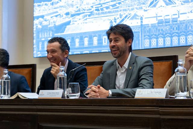 Dos hombres sonriendo durante un evento en una sala con fondo arquitectónico.
