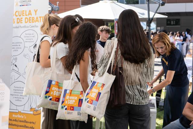 Un grupo de personas observa un stand de información al aire libre con bolsas de tela en un evento educativo.