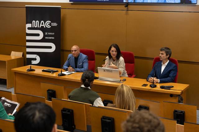 Una presentación en una sala con tres personas sentadas delante de un micrófono y un logotipo visible en la pared.