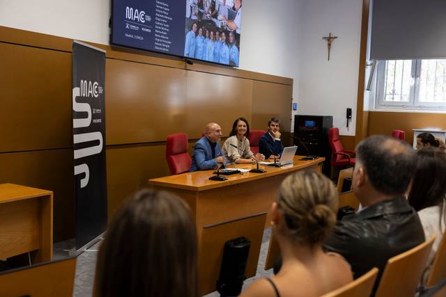 Una presentación en una sala con tres ponentes hablando frente a una audiencia sentada.