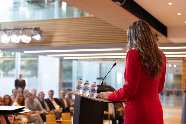 Una mujer de espaldas con un abrigo rojo habla en un podio frente a una audiencia sentada.
