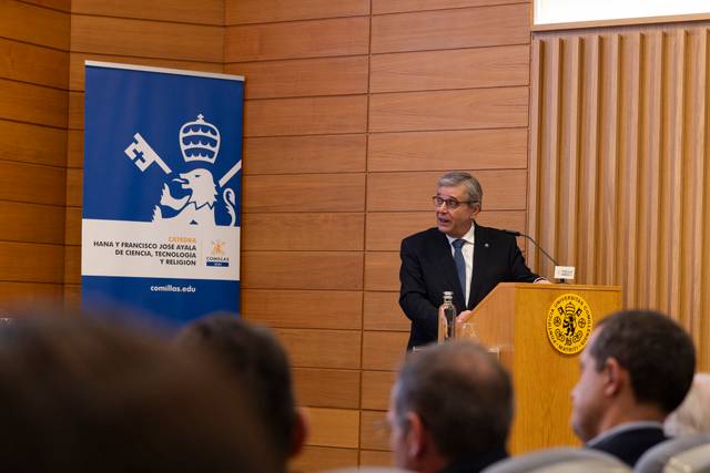 Un hombre presenta en un evento formal junto a un banner institucional.
