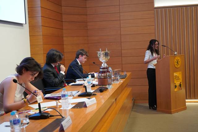 Una presentadora habla en un evento mientras un grupo de personas la escucha atentamente.