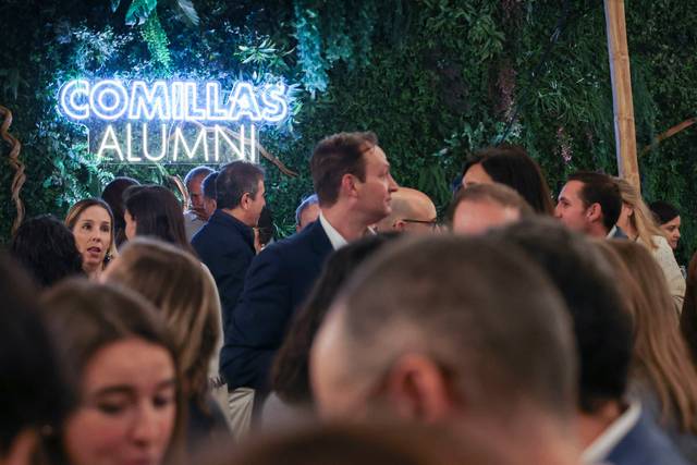 Un evento social con muchas personas conversando bajo un letrero luminoso que dice 'COMILLAS ALUMNI'.