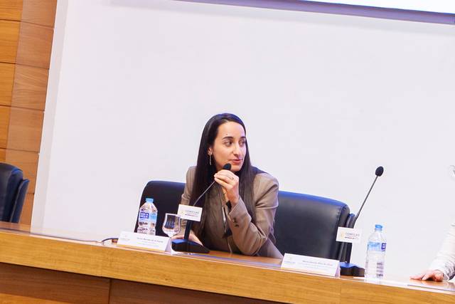 Una mujer sentada en una mesa durante un evento o conferencia, sosteniendo un micrófono y mirando hacia un lado.