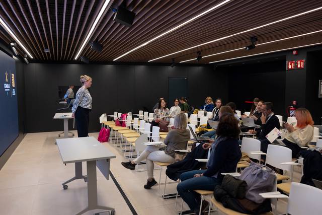 Una sala de conferencias con una presentadora ante un público atento.