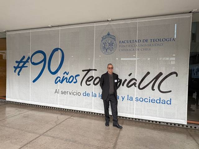 Un hombre posando frente a un mural que celebra los 90 años de la Facultad de Teología de la Pontificia Universidad Católica de Chile.