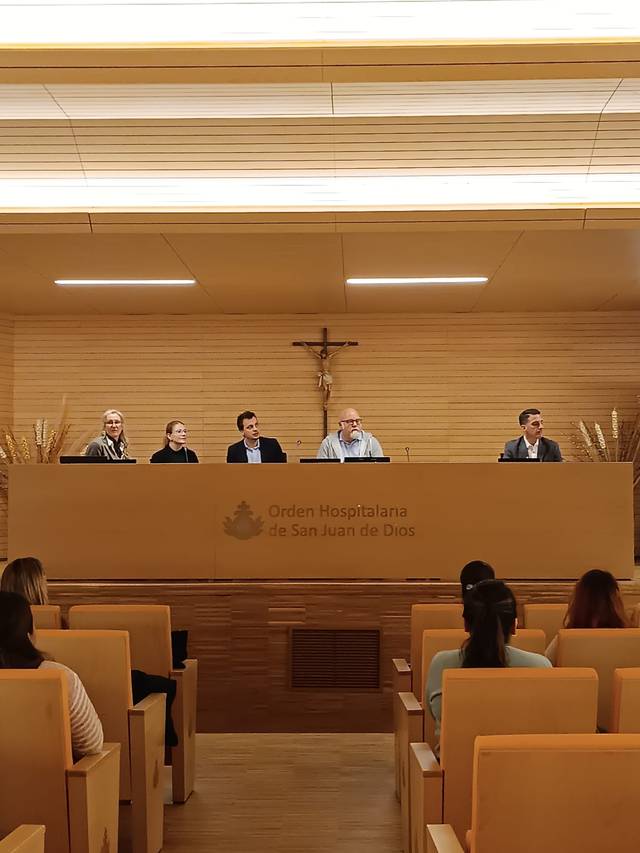 Cinco personas sentadas en un panel de conferencias dentro de una sala con audiencia y un crucifijo en la pared.