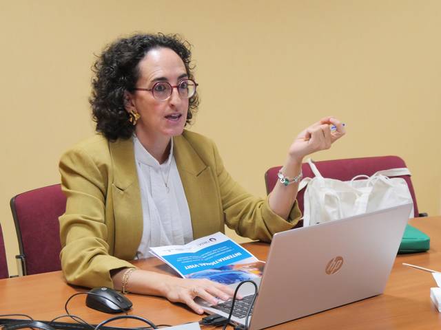 Una mujer con cabello rizado y gafas está dando una presentación en una reunión mientras señala un documento sobre la mesa.