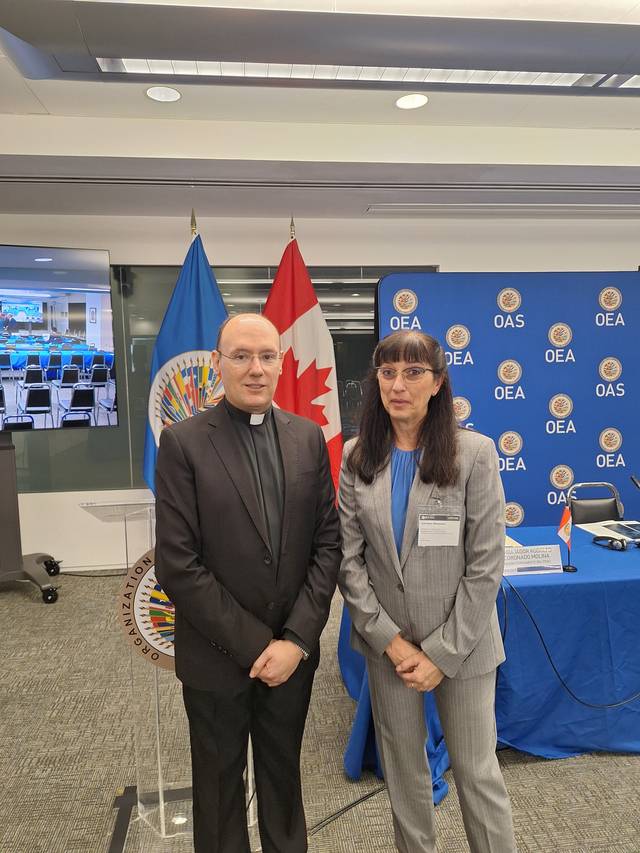 En la imagen se encuentran dos personas posando frente a banderas, una de Canadá y otra de la OEA.