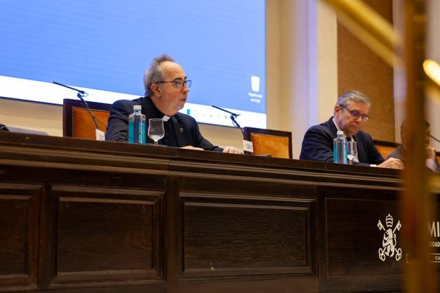 Dos hombres sentados en una mesa larga y oscura en lo que parece ser un evento o conferencia dentro de una sala elegante.