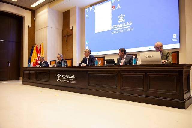 A panel of four men seated at a long table in a conference hall discussing, with a projection screen displaying a university logo in the background.