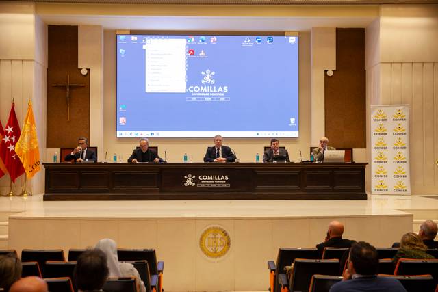 Panel de conferencia en una universidad con cinco personas sentadas detrás de una mesa y público presente.