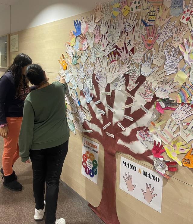 Dos personas observan un mural con forma de árbol hecho de manos de papel con dibujos y mensajes.