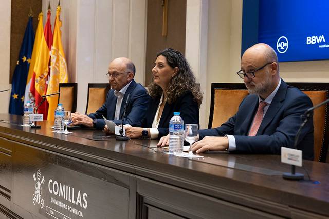 Un grupo de tres personas sentadas frente a un panel en la Universidad Pontificia Comillas, con banderas de España al fondo.