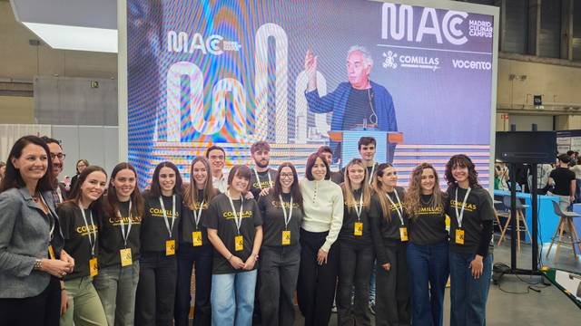 Un grupo de personas posando frente a una pantalla en un evento con un ponente en el fondo.