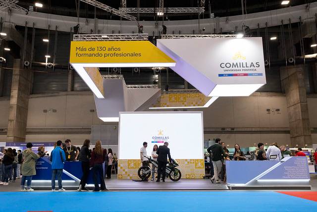 Un stand de la Universidad Pontificia Comillas en una feria, con un gran cartel que destaca sus más de 130 años formando profesionales.