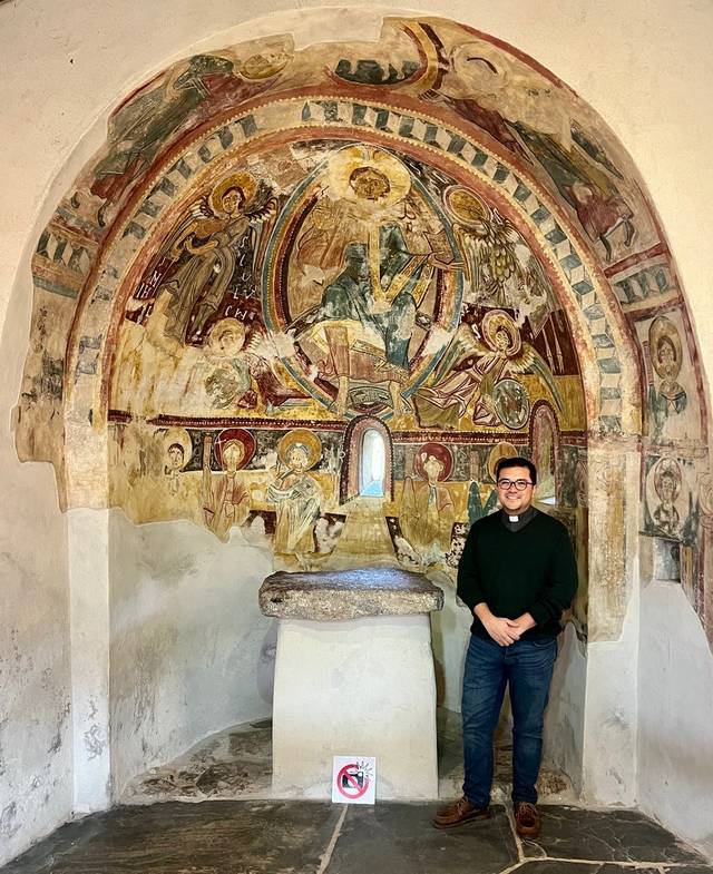 Un hombre sonriente posa frente a una pared con pinturas religiosas en una capilla antigua.