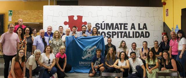 Un grupo grande de personas posando frente a un mural que dice 'SÚMATE A LA HOSPITALIDAD'.