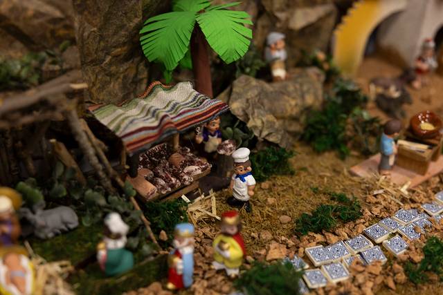 La imagen muestra un belén con figuras en un paisaje rural, destacando un puesto de comida y un fondo decorado con vegetación.