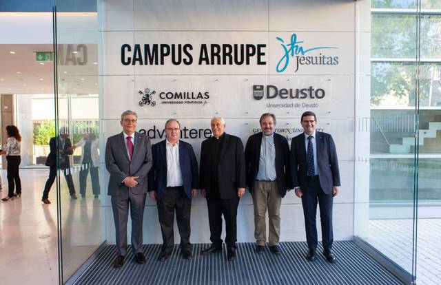 Five men standing in front of the entrance of Campus Arrupe, with signs of Jesuit universities Comillas and Deusto.