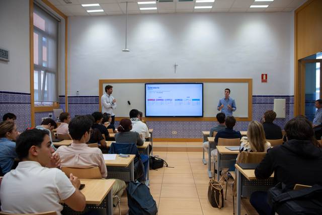 Una clase donde dos presentadores exponen frente a una audiencia de estudiantes en un aula moderna.