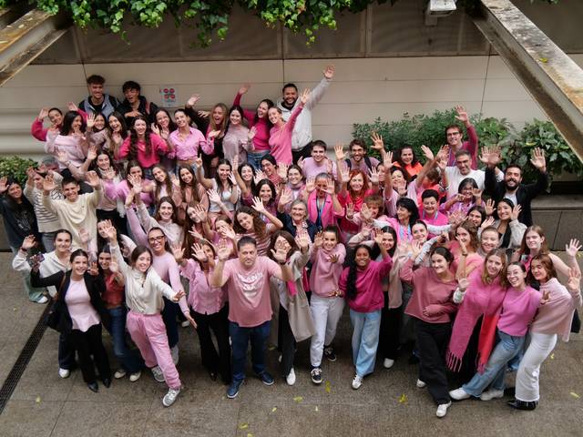 Un grupo grande de personas sonrientes levantando las manos en un ambiente al aire libre, todas vestidas en tonos rosa.