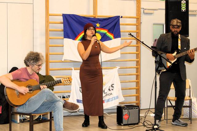 Un grupo musical se presenta en un escenario mientras una mujer canta y dos músicos la acompañan con guitarra y bajo.
