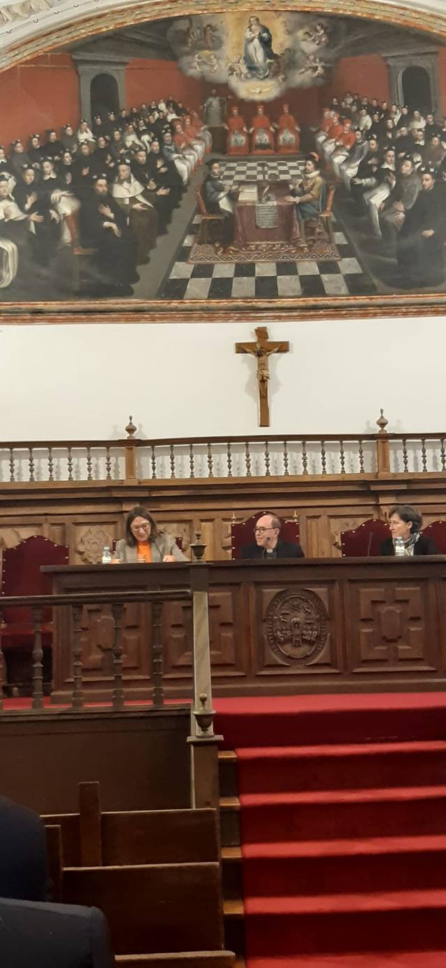 Three people are sitting at a panel in a historical room with a large painting in the background and a crucifix above them.