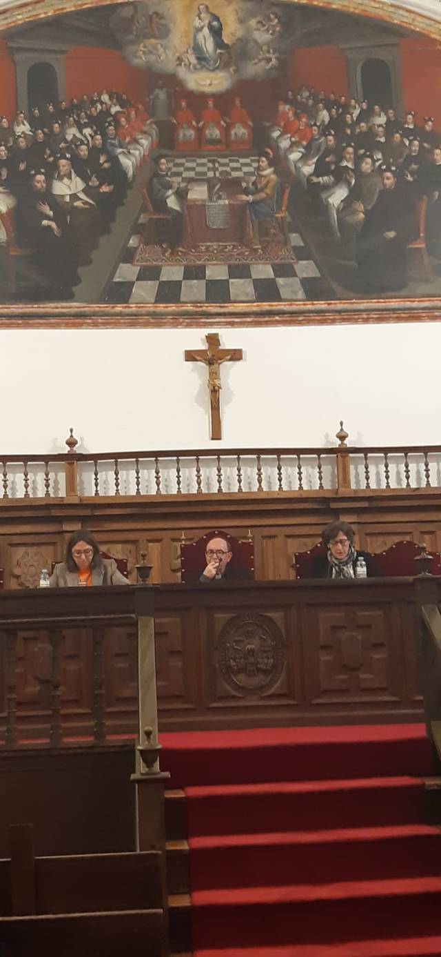 An image showing people sitting in an ornate wooden courtroom or assembly hall, with a large historical painting in the background, a crucifix at the center, and red carpeted stairs in the forefront.