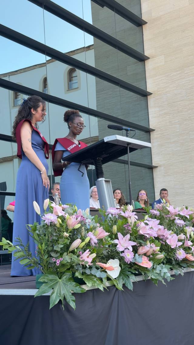 Una ceremonia con dos oradoras en un escenario adornado con flores, frente a un grupo de asistentes.