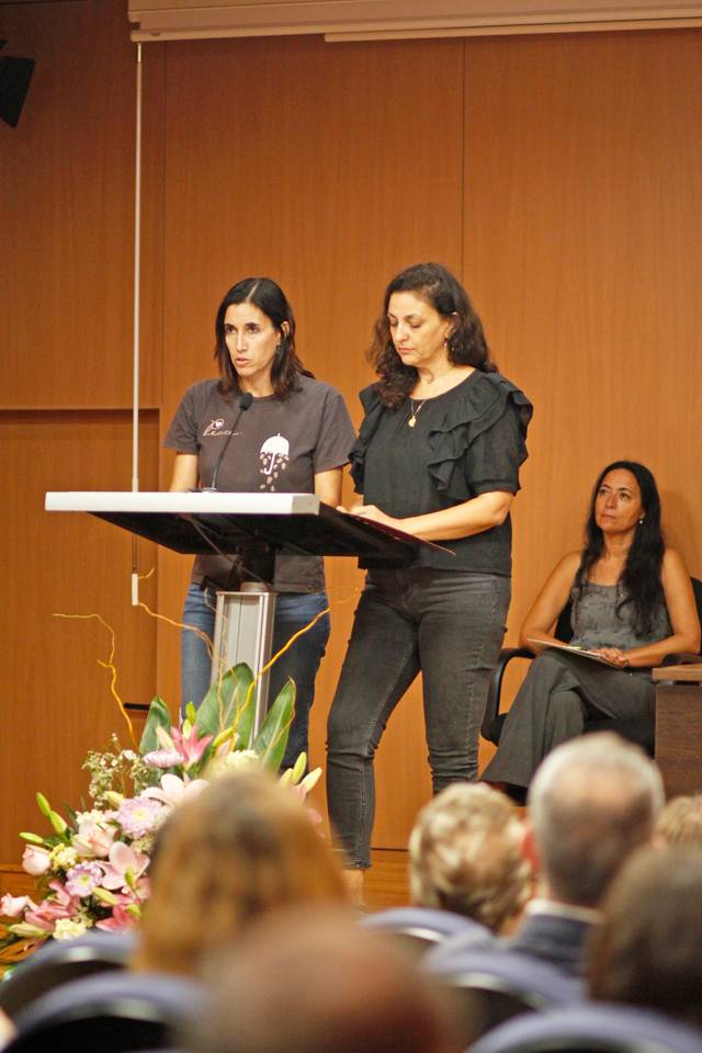 Two women standing at a podium in a conference room with an audience, with another woman seated in the background.