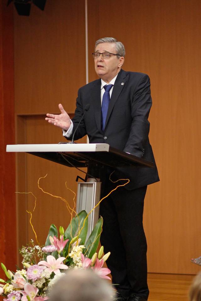 A man in a suit and glasses speaking at a podium with floral decorations in a conference room.