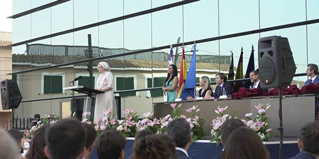 A person in a white robe is giving a speech at an outdoor graduation ceremony in front of a seated audience, with officials and flags in the background.