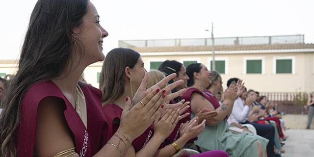 A group of women, dressed in maroon sashes, clapping hands while sitting in an outdoor event.