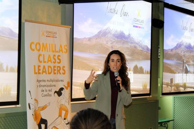 Una presentadora habla frente a una audiencia en un evento, con un paisaje montañoso proyectado de fondo.