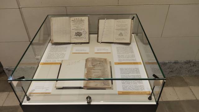 La imagen muestra una vitrina con varios libros antiguos y documentos, acompañados de etiquetas informativas.