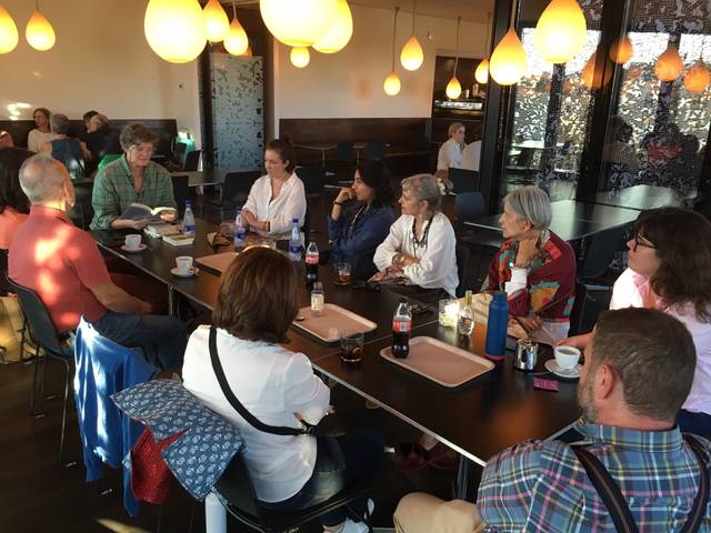Un grupo de personas sentadas en una mesa en un café, conversando y prestando atención a una mujer que lee.