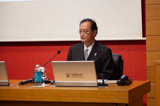 Un hombre de mediana edad con gafas viste un traje oscuro y está sentado frente a una computadora en un auditorio con el logo de Comillas visible.