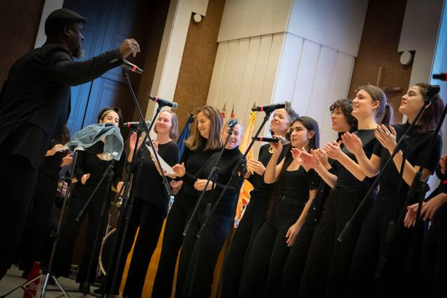 Un director de coro afroamericano dirigiendo a un grupo de cantantes femeninas durante una actuación.