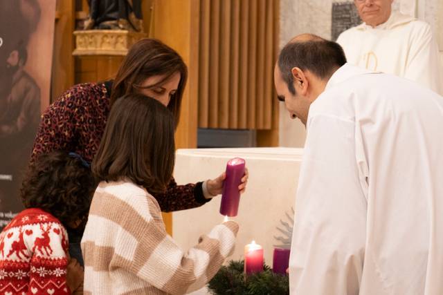 Una mujer y una niña encienden una vela mientras un hombre observa en un ambiente religioso, con velas y un sacerdote de fondo.