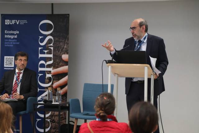 A man is giving a speech at a lectern during a conference, with another man seated beside him and an audience in front.